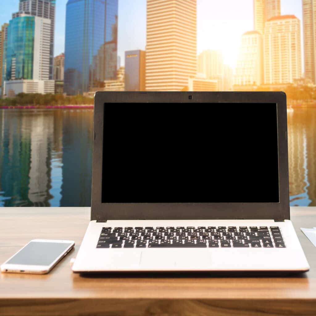 Mockup image of laptop with blank white screen on wooden table view outdoors of office building cityscape background.