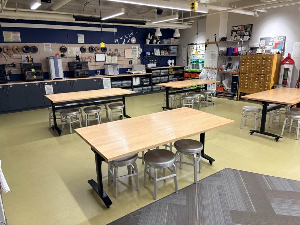A room with several tables, stools, and 3D printers across the back wall.